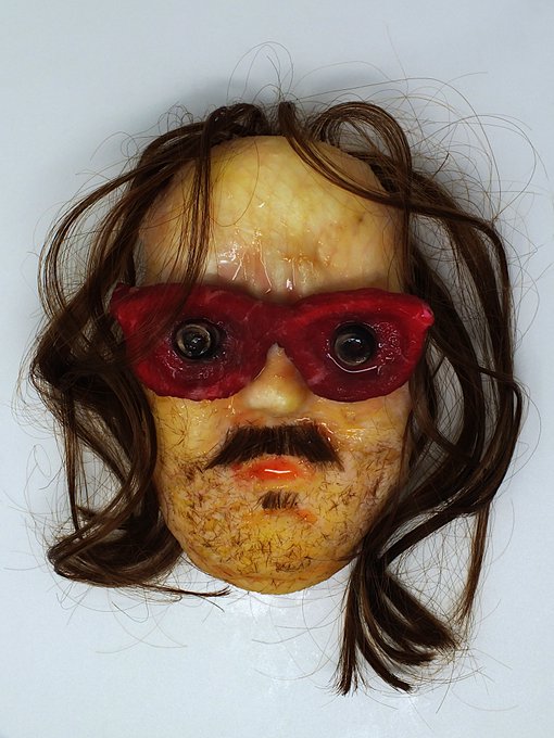 A creepy handmade mask featuring real human hair, mustache, red lips, and large eyes with red frames, photographed against a white background.