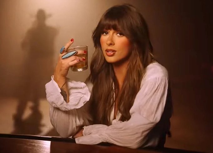 A woman with long brown hair and bangs, wearing a white blouse, holds up an empty glass while wearing a large turquoise ring, with a cowboy-hat-shaped shadow cast on the wall behind her.