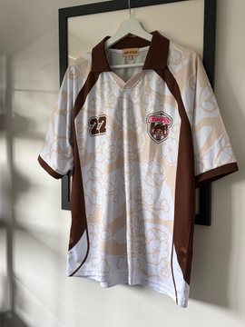 A white and brown vintage-style sports jersey with the number 22 and a circular emblem hangs on a hanger against a white wall.