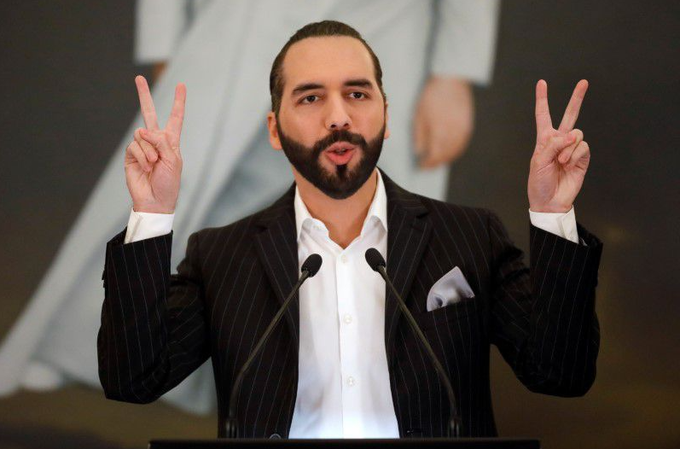A man in a pinstripe suit and white shirt standing at a podium, holding up both hands in peace signs.