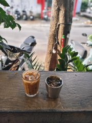 Two glasses of coffee sit on a wooden ledge outdoors, with a glass bottle, plants, and motorbikes visible in the background.