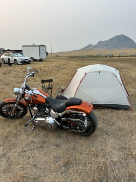 A scenic outdoor photograph of a campsite featuring an orange motorcycle parked in front of a gray and orange tent, with mountains and RVs/trailers visible in the background on dry grassland.