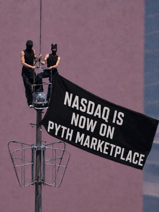 Two masked individuals sit atop a pole displaying a banner that reads 'NASDAQ IS NOW ON PYTH MARKETPLACE'.