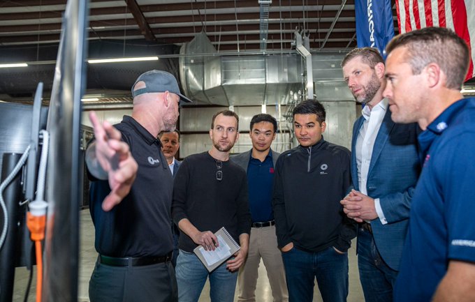 The image shows a group of six men gathered around a table or workbench inside a building with a large American flag hanging from the ceiling. One man appears to be giving instructions, possibly related to Bitcoin, while the others listen attentively. The scene suggests that these individuals are engaged in a discussion about Bitcoin and its potential applications or implications for their business or industry.