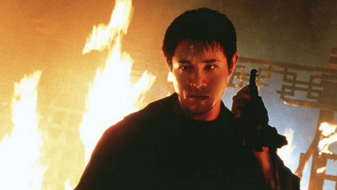 A man holding a gun stands in front of a blazing fire in a dramatic, warmly lit scene.