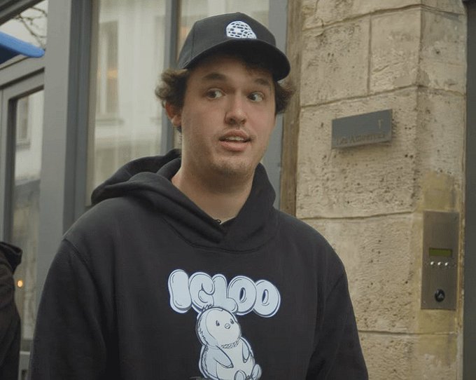 A young man wearing a black cap and a black hoodie with an 'IGLOO' graphic featuring a dinosaur, standing outside near a stone building.