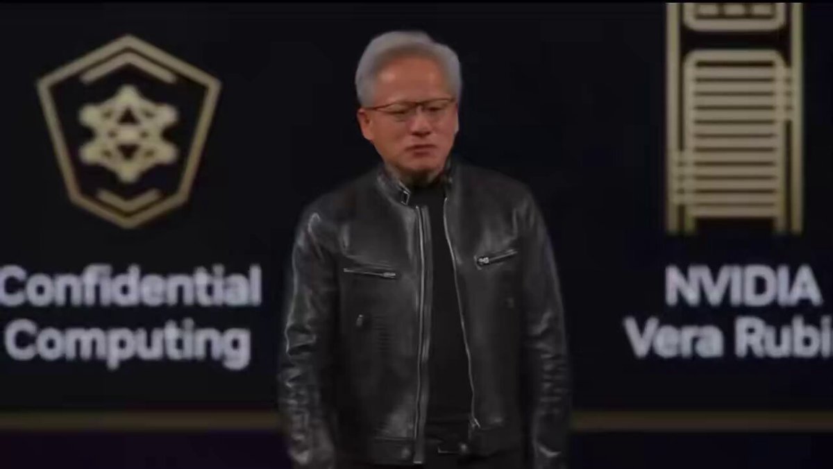 The image features an older man wearing a black leather jacket, standing in front of two logos - one representing Confidential Computing byNVDA and another representing Vera Rubi by nebius. The man appears to be giving a speech or presentation on stage, with his hands placed behind him as he speaks.