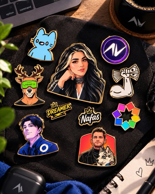 A collection of enamel pins displayed on a dark surface, featuring a blue cat, a deer with 'ARMETTA' on its glasses, a woman with black and white hair, an 'AV' logo, a flexing arm, text reading 'DREAMER' and 'Nafas', a color wheel, a man in a blue suit, and a man with a wolf dog.