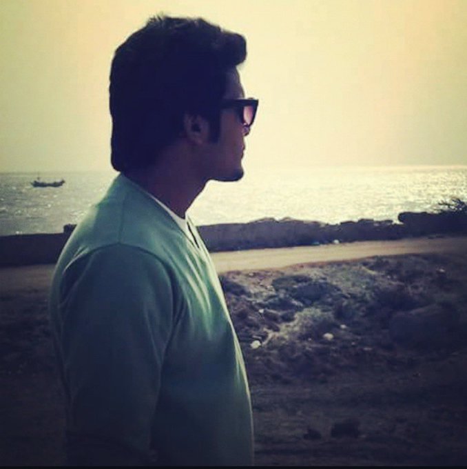 A man with dark hair, sunglasses, and a goatee stands in profile wearing a teal t-shirt, gazing out at a calm sea at sunset.