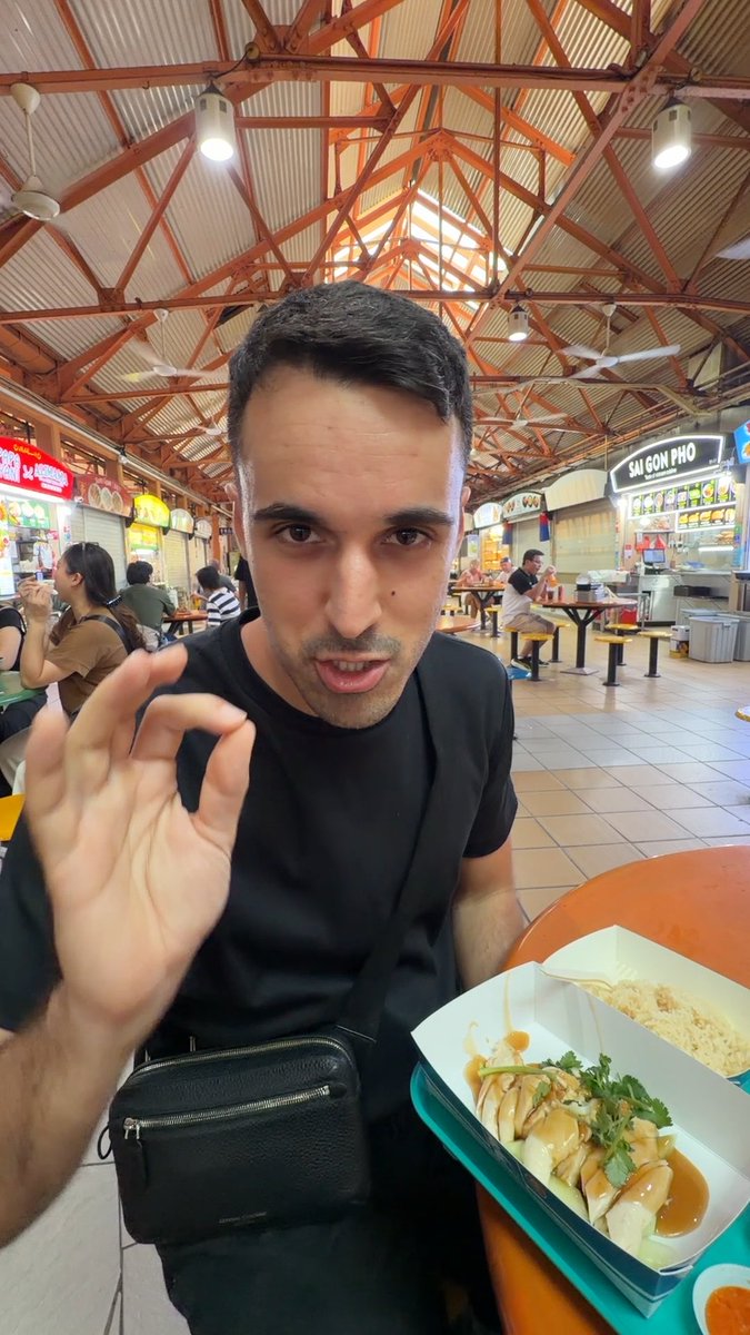 A man in a black shirt makes an OK gesture while sitting at an outdoor hawker center, with a tray of food (rice and meat with sauce) in front of him.