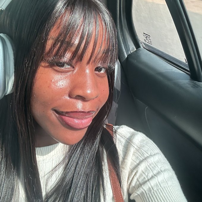 A close-up selfie of a woman with long dark hair and bangs sitting in the driver's seat of a car.