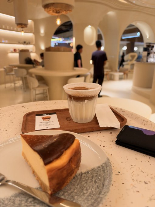 A slice of Basque burnt cheesecake on a plate and a cup of coffee on a wooden tray in a modern, stylish café with arched doorways and a terrazzo table, with people visible in the background.