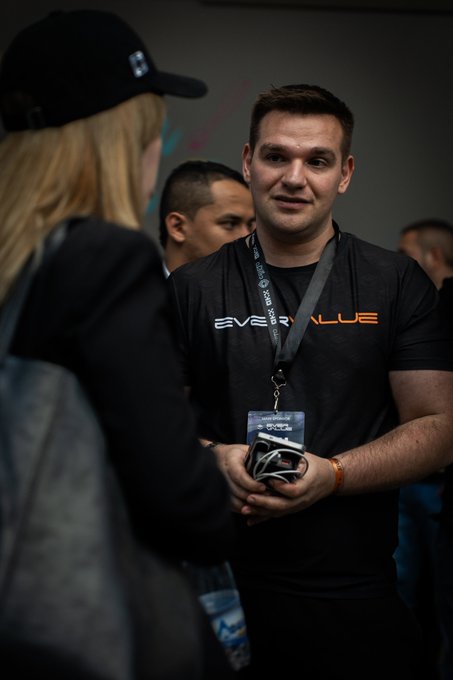 A man wearing an "EVERVALUE" t-shirt and a lanyard converses with a blonde woman in a black cap at what appears to be a tech or networking event.