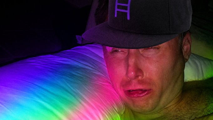 A person wearing a black cap with a purple "H" logo appears emotional, illuminated by rainbow-colored lighting in a darkened setting.