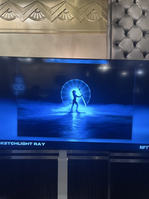 A TV screen displaying a blue 'SKETCLIGHT RAY' NFT artwork featuring a figure with a glowing orb, set against a tufted wall background.