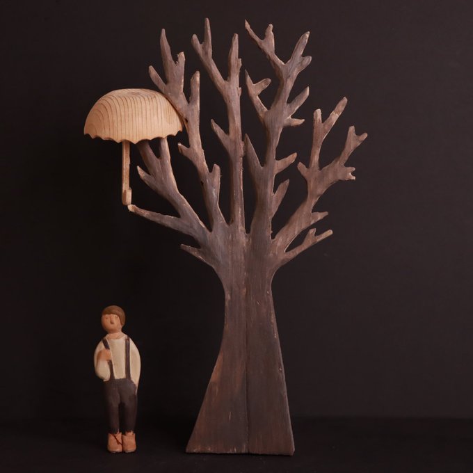 A small wooden figurine stands beside a dark, bare tree sculpture, where a light-colored umbrella appears to have caught on the branches, perfectly complementing the solitary, whimsical charm of "a good lone tree."