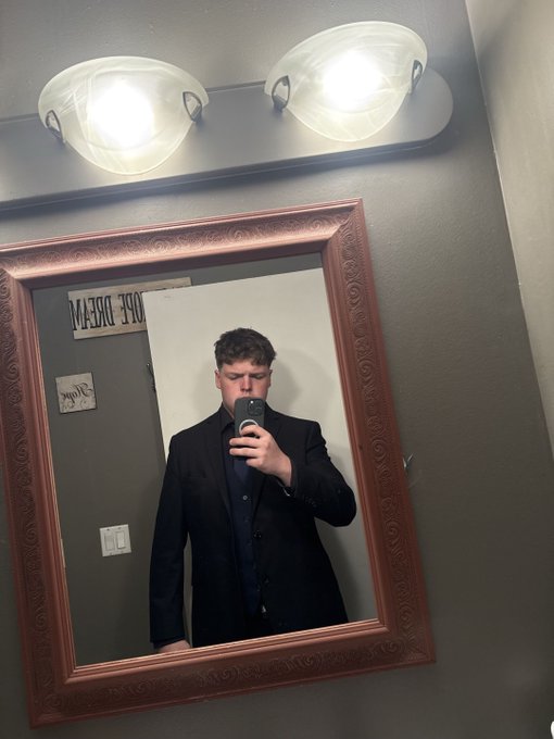 A young man in a black suit takes a mirror selfie with his phone, standing under a lit vanity mirror in a bathroom, with reversed text 'TOE' and 'M' visible on the wall behind him.