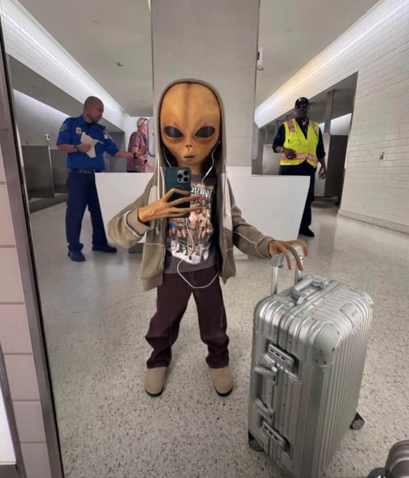 A person wearing an alien mask and gray hoodie takes a mirror selfie in what appears to be an airport or transit hub, pulling a large silver Rimowa suitcase while a TSA officer and someone in a high-visibility vest stand in the background. The image humorously embodies the post's theme of embracing an otherworldly, chaotic "CT" (crypto Twitter) lifestyle—suggesting the alien-like absurdity of diving headfirst into crypto culture after just opening one account.