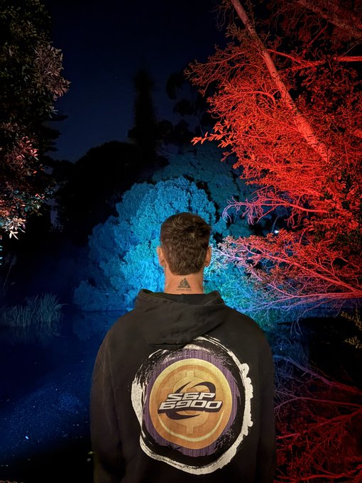 A nighttime photo of a person from behind wearing a black hoodie with 'LOS SISHU' (or similar) logo, standing before trees illuminated by red and blue lighting.