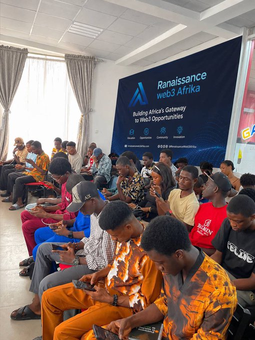A group of people sit on benches looking at their phones in a room with a large 'Ranaissance web3 Afrika' banner that reads 'Building Africa's Gateway to Web3 Opportunities'.