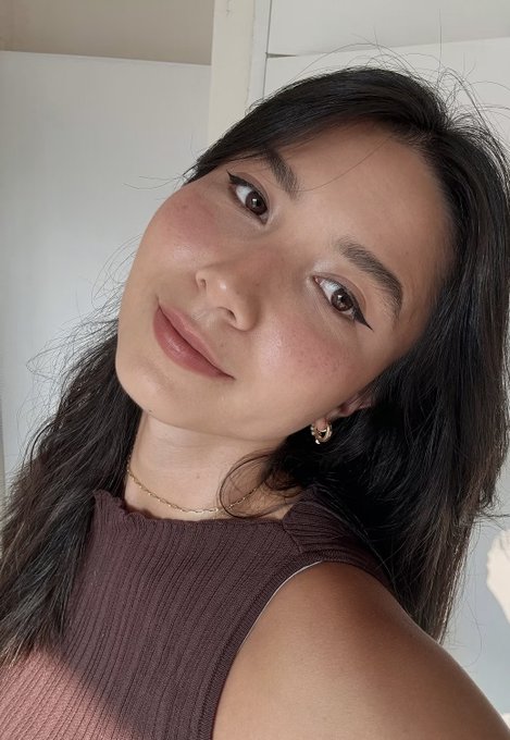 A selfie of a young woman with long dark hair wearing a brown sleeveless top, small gold hoop earrings, and a delicate gold necklace, with soft natural lighting.