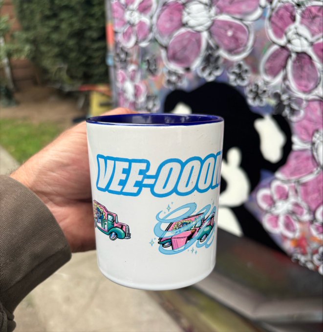A hand holding a white enamel mug with a blue rim featuring 'VEE-OOO' text and colorful, stylized vehicle graphics, set against a graffiti-covered background.