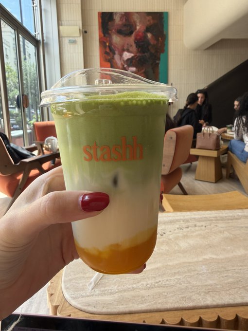 A hand with red nail polish holds a clear plastic cup branded 'stashh' containing a layered matcha drink with mango at the bottom, photographed in what appears to be a café setting.