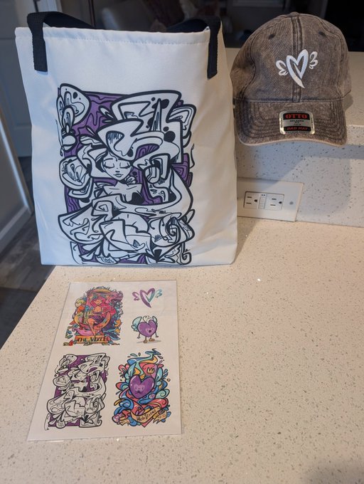 A display of merchandise on a kitchen counter featuring a white tote bag with a purple artistic print, a brown trucker hat with a heart logo, and a sheet of colorful stickers with similar artistic designs.