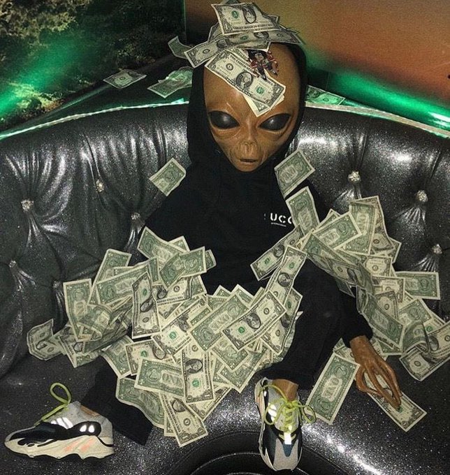 A person wearing an alien mask and black hoodie sits on a tufted leather couch, covered in scattered US dollar bills, wearing white and black sneakers.