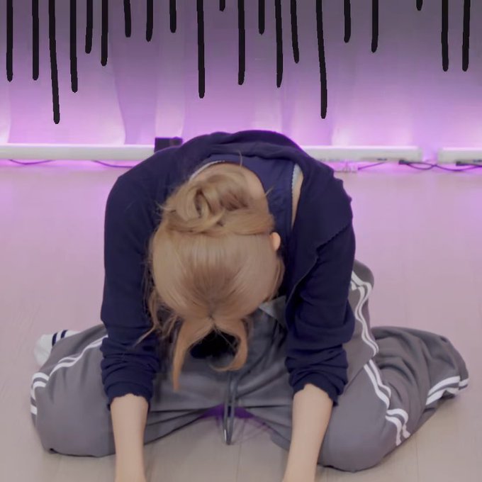 A blonde-haired person in a dark hoodie and grey track pants sits on the floor bowing forward with their head down against a purple backdrop with black drip decorations.