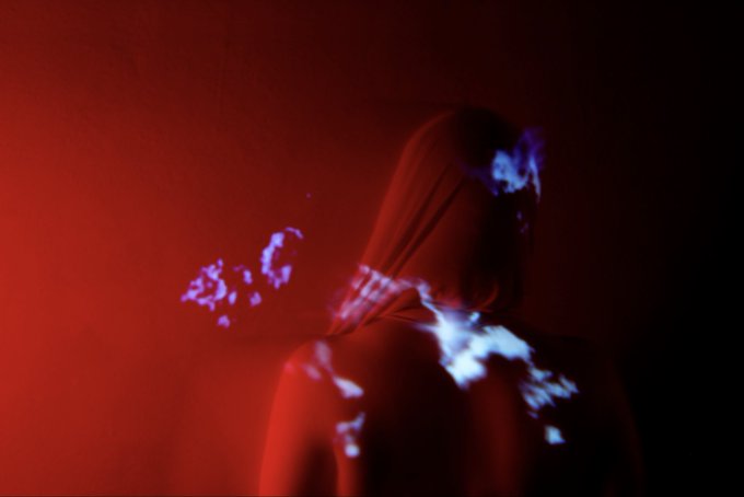 A hooded figure stands against a dark red background with blue glowing particle effects emanating from their body.