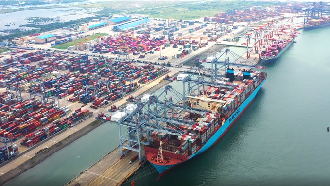Aerial view of a busy commercial port featuring a large Maersk container ship docked alongside rows of stacked shipping containers and gantry cranes.