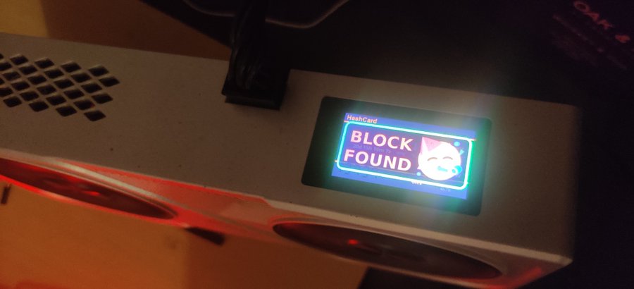 A close-up of a Bitcoin mining machine displaying a lit screen that reads 'HashCard BLOCK FOUND' next to a smiling cat logo, with the device's fans glowing red.