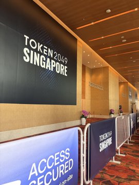 Photo of the Token2049 Singapore conference entrance, showing a large black sign with white text 'TOKEN 2049 SINGAPORE' and blue barriers reading 'ACCESS SECURED'.