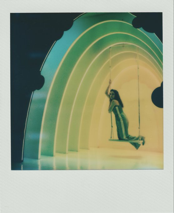 The image features a woman sitting on a swing suspended from an archway, which has a rainbow-colored design. She appears to be enjoying her time swinging through the air. The photo is labeled with the hashtag #Polaroid, indicating that it was taken using a Polaroid camera.