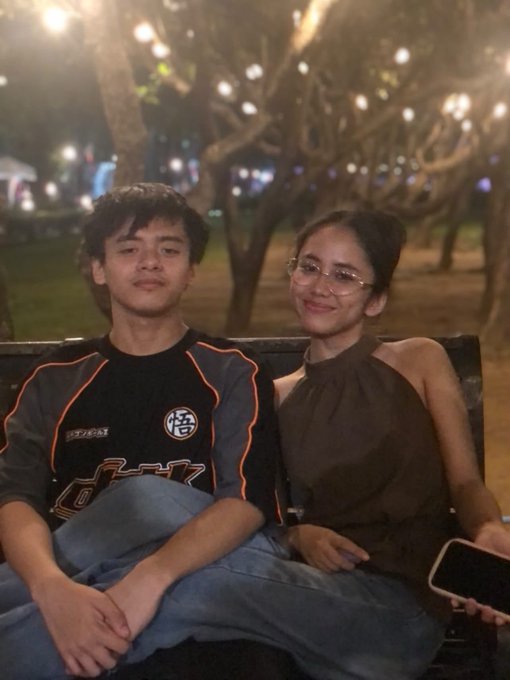 A young man in a Dragon Ball themed shirt and a young woman in a brown top and glasses sit together on a bench at night with illuminated trees in the background.