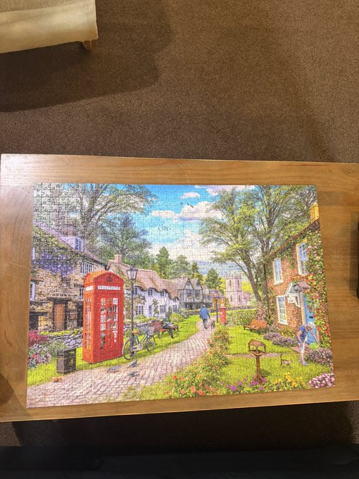 A completed jigsaw puzzle of an English village scene with thatched cottages, a red telephone box, and flowers, resting on a wooden table.