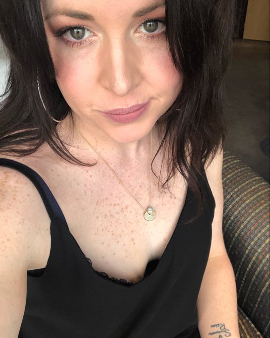 A woman with dark hair takes a selfie, showing off a necklace and a tattoo on her arm.