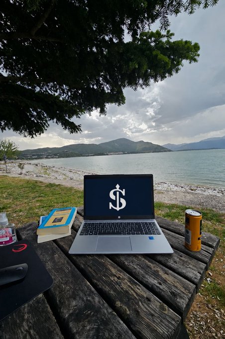 A laptop displaying a large white dollar sign on its screen sits open on a wooden picnic table by a serene lake, accompanied by a stack of books, a mouse, and a canned beverage.