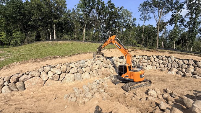 An orange Doosan excavator is shown constructing or arranging a stone wall in a wooded area. The accompanying post sarcastically mocks Bitcoin security influencers who apparently got their Coldcards compromised, suggesting basic security practices ("the assignment") were neglected in favor of trivial proof-of-work.