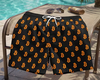 A pair of black swim shorts with an orange Bitcoin 'B' pattern, displayed on a chair by a pool with sunglasses and sunscreen nearby.