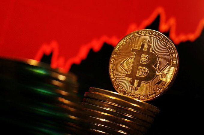 A gold Bitcoin coin stands upright on a stack of coins against a red background featuring a downward-trending line graph.