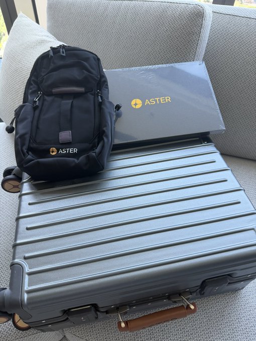 A black Aster-branded backpack sits atop a sleek gray hard-shell suitcase, both featuring the gold Aster logo, placed on a light-colored couch. The image showcases the merchandise received from Aster DEX, as mentioned in the post thanking the platform and Ccweb3hub for the gifts.