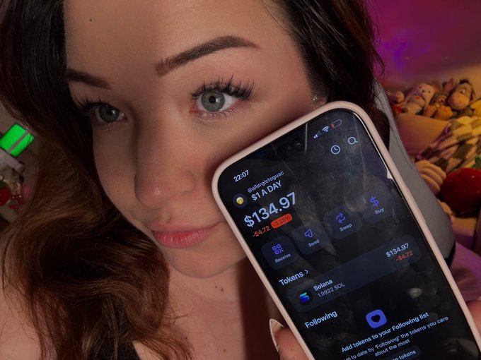 In the image, a woman with brown hair and green eyes is holding up her cell phone displaying various information about her transaction. The main text she sees is "day 198 of buying $1 worth of sol until atg's". This could possibly be related to the stock market or an online shopping website where she has recently made a purchase. She might be discussing this situation with someone, as suggested by another post that says "let me know about this".