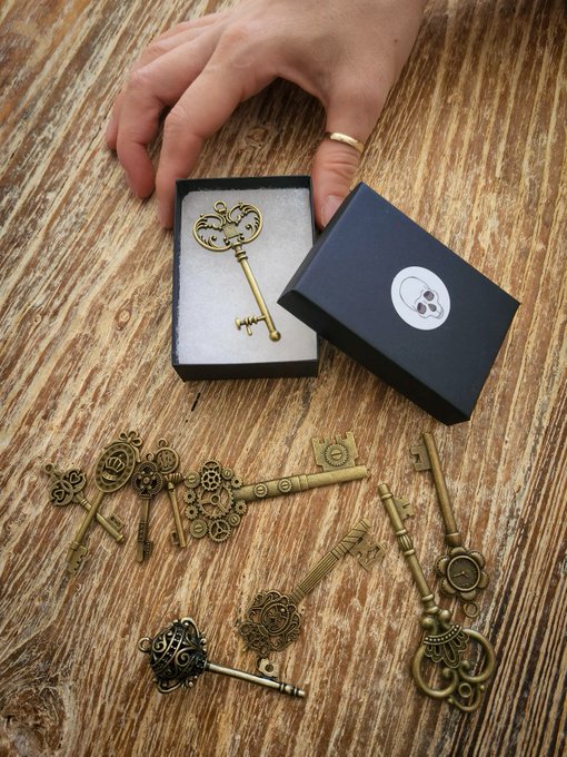 A hand holding a small black gift box displaying an ornate gold mouse-shaped key pin, with a collection of other antique-style brass skeleton keys scattered on a wooden surface below.