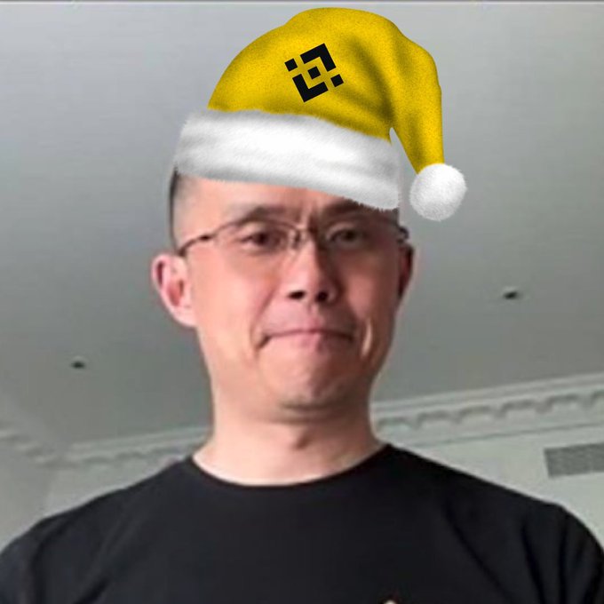 A close-up of a man with a yellow Santa hat digitally edited onto his head, featuring a black logo resembling the Binance symbol.