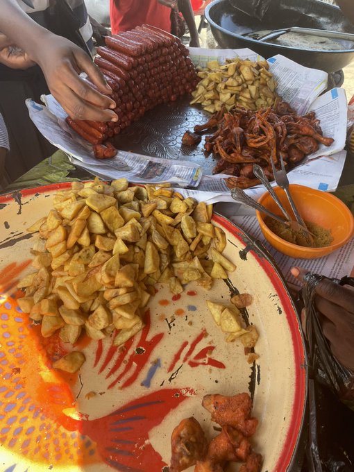 A street food scene showing a vendor's hands handling a tray of bright red sausages alongside piles of fried fritters and reddish snacks on platters lined with newspaper.
