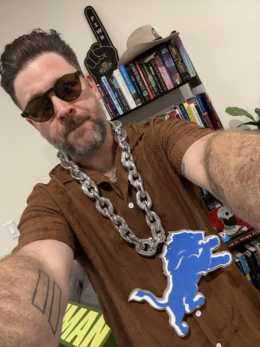 A selfie of a bearded man wearing sunglasses and a large silver chain necklace, with a blue Detroit Lions logo sticker overlaid on his brown shirt, taken in front of a bookshelf.
