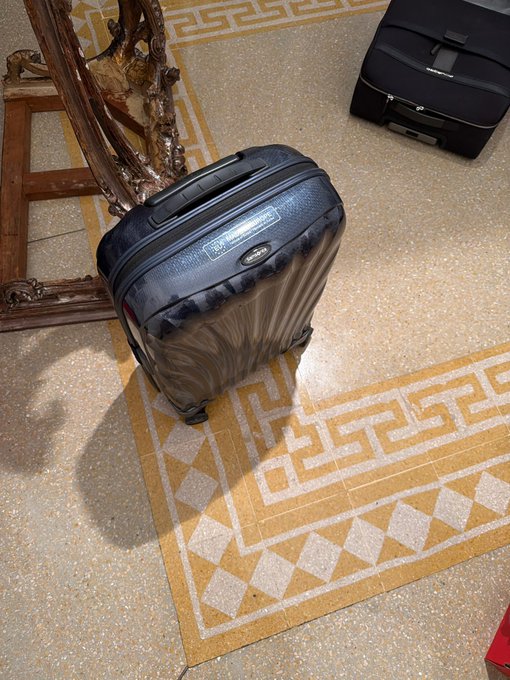 Two dark-colored hardshell rolling suitcases sit on a tiled floor featuring a geometric Greek key pattern border, near an ornate carved wooden chair leg.