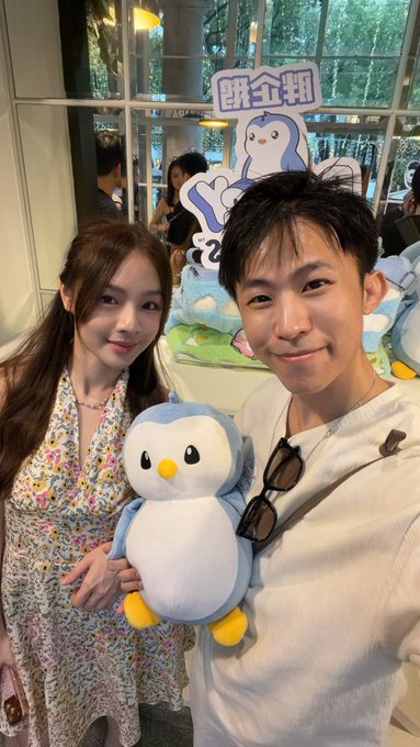 A couple poses at the Pudgy Penguins event, smiling as the woman holds a plush penguin toy, with a cartoon penguin display behind them reading "周企划" (Week Plan), celebrating the brand's ongoing legitimacy in contrast to rug-pulled NFT projects.